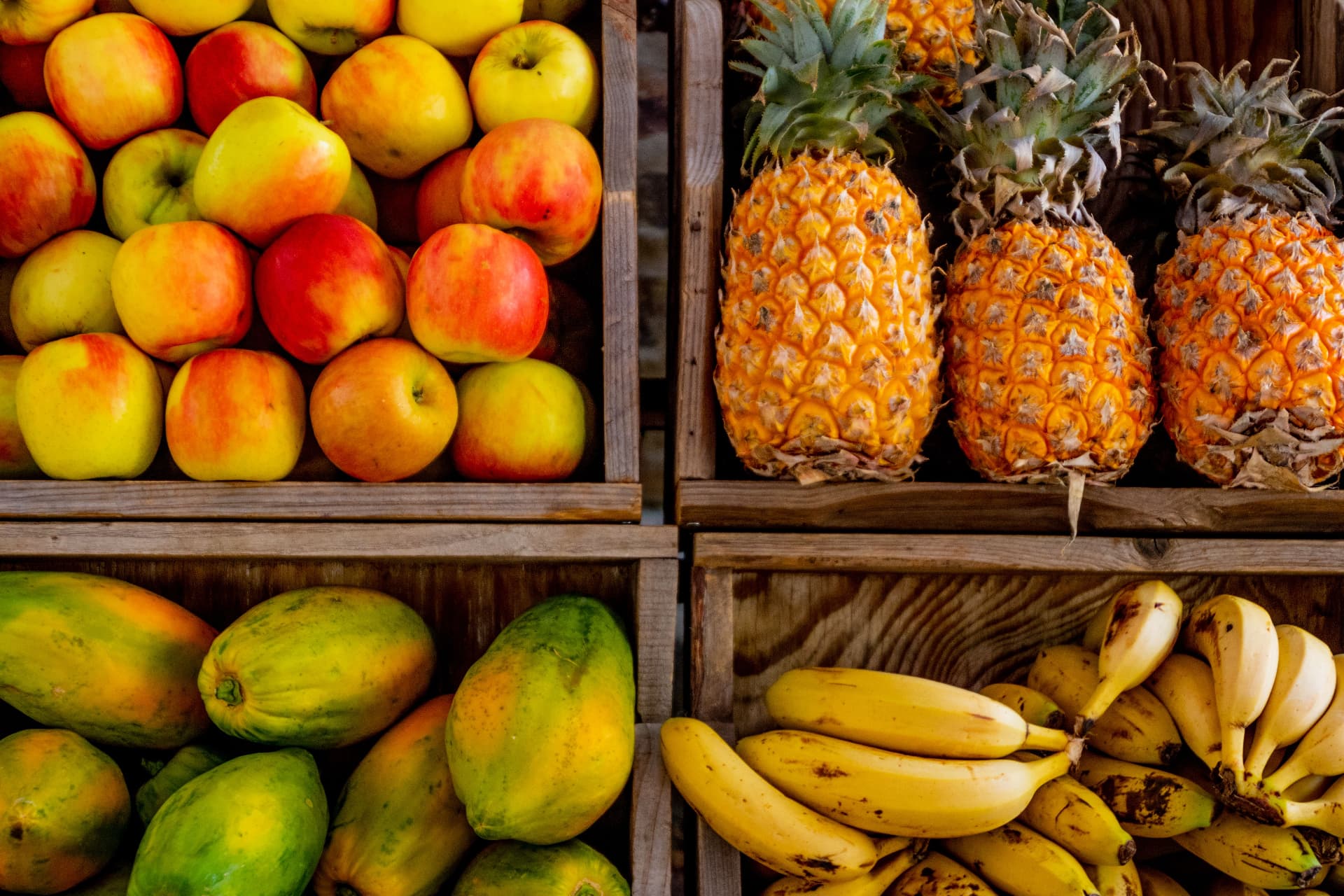 Holzkisten mit rotgelben Äpfeln, Ananas, grün-gelben Papayas und Bananenbüscheln übereinander gestapelt